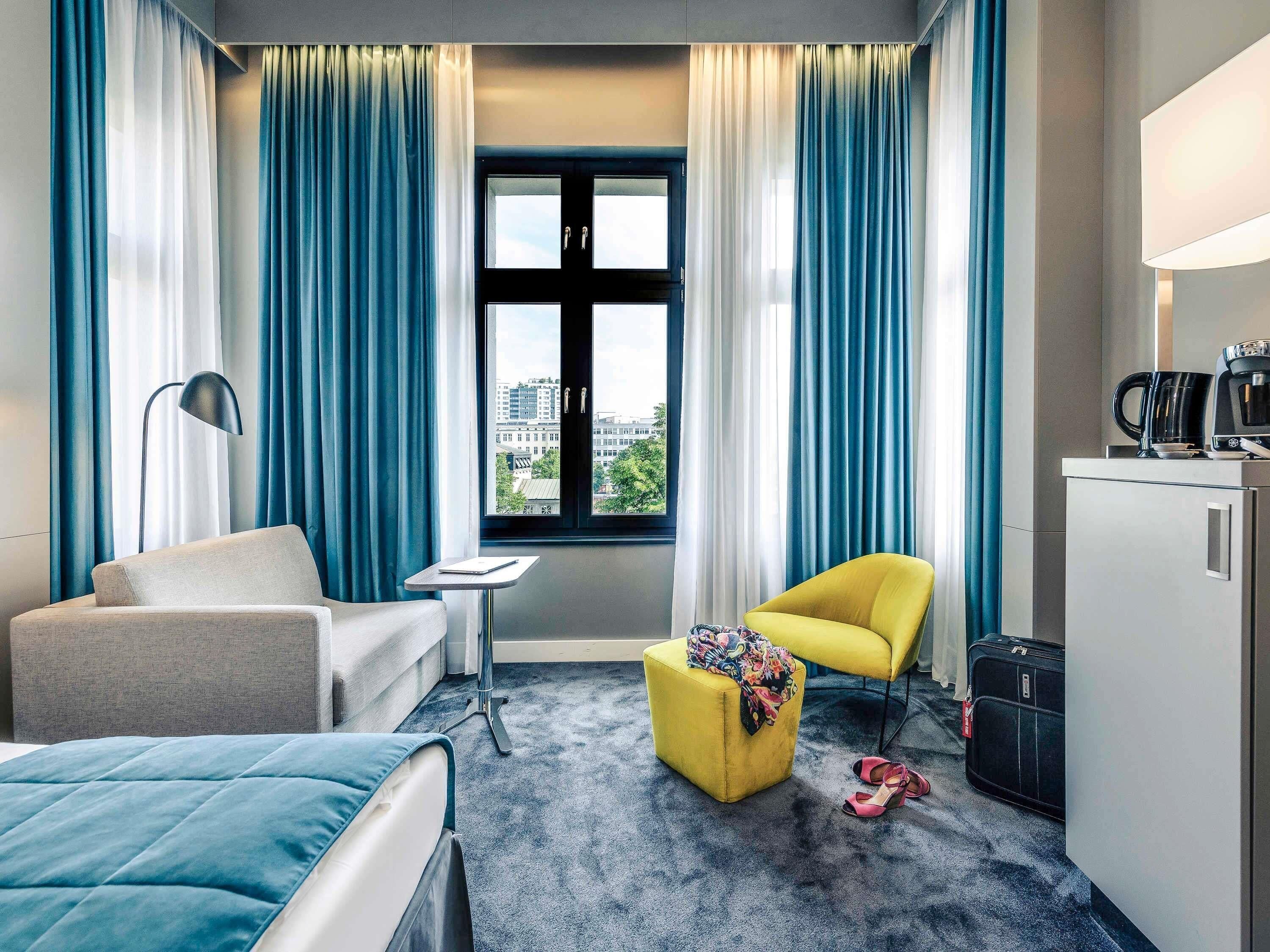 Ein Hotelzimmer mit einem Bett, einem Schlafsofa, einem gelben Sessel, einem Schreibtisch, einer Lampe, einer Kaffeemaschine und Blick auf die Stadt durch ein Fenster.