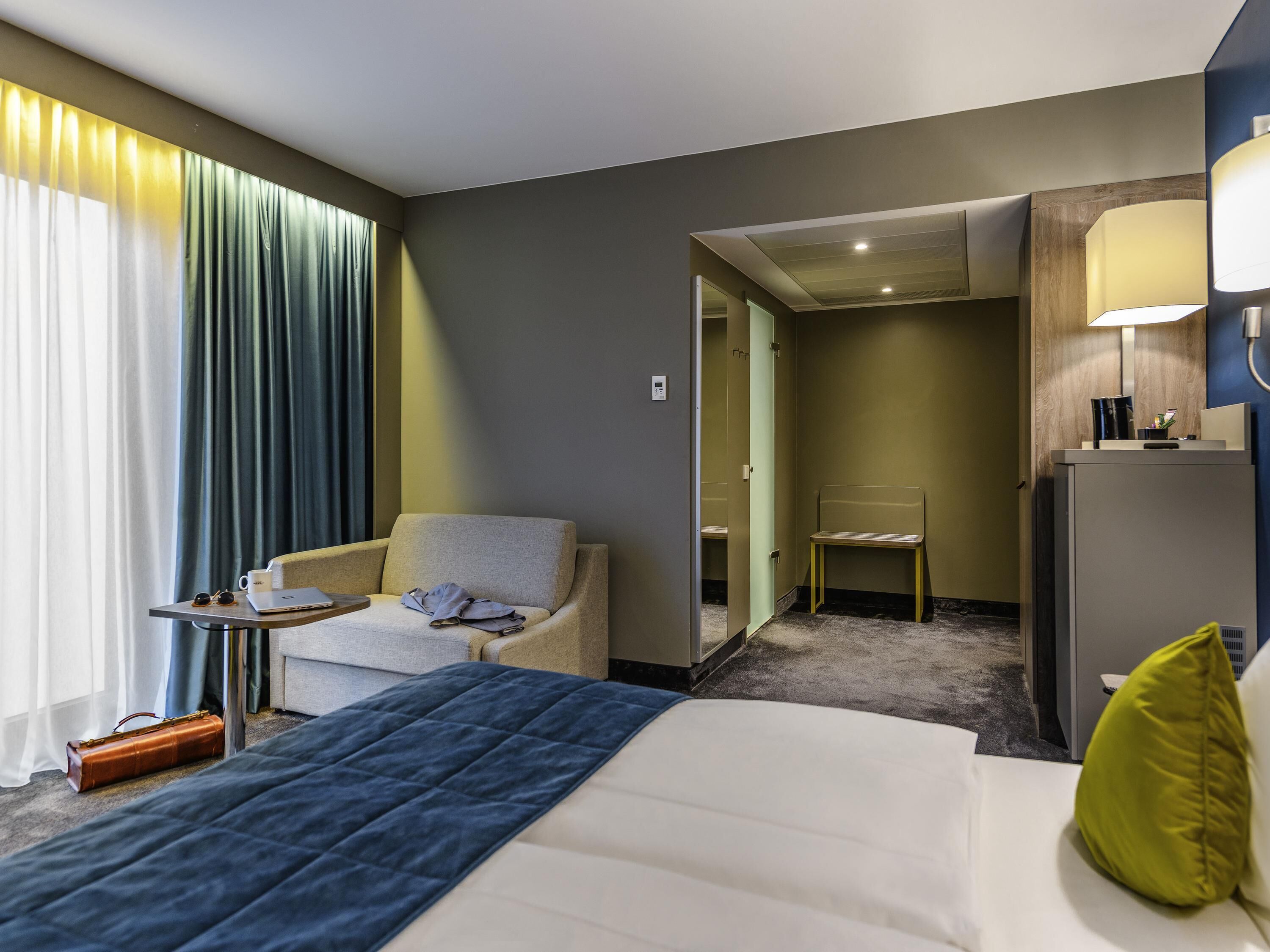 Habitación de hotel con una cama, una silla, una mesita con una computadora portátil y vasos, y un escritorio con una lámpara.