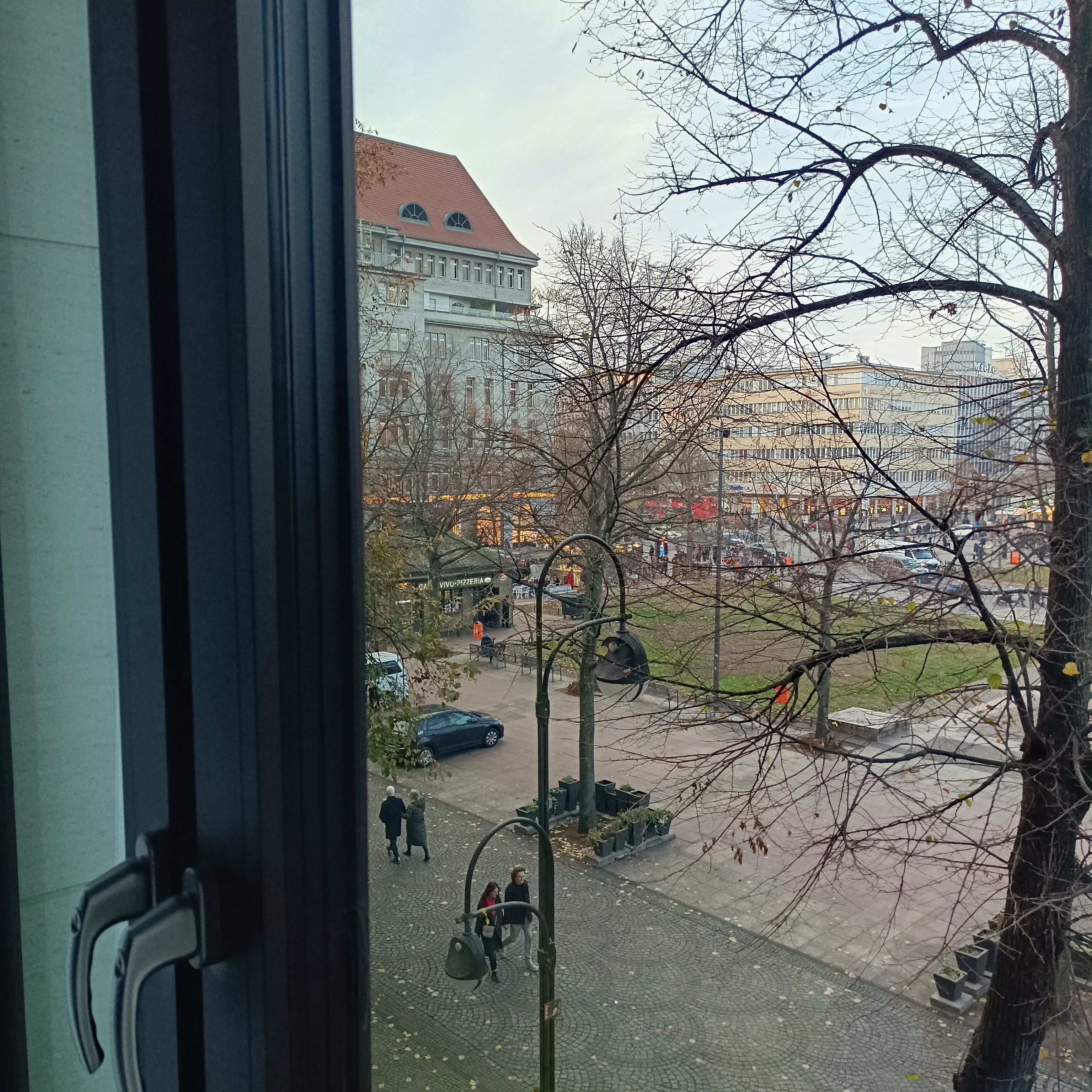 Vista desde el interior de un edificio, mostrando una calle con edificios, un árbol y algunas personas caminando.