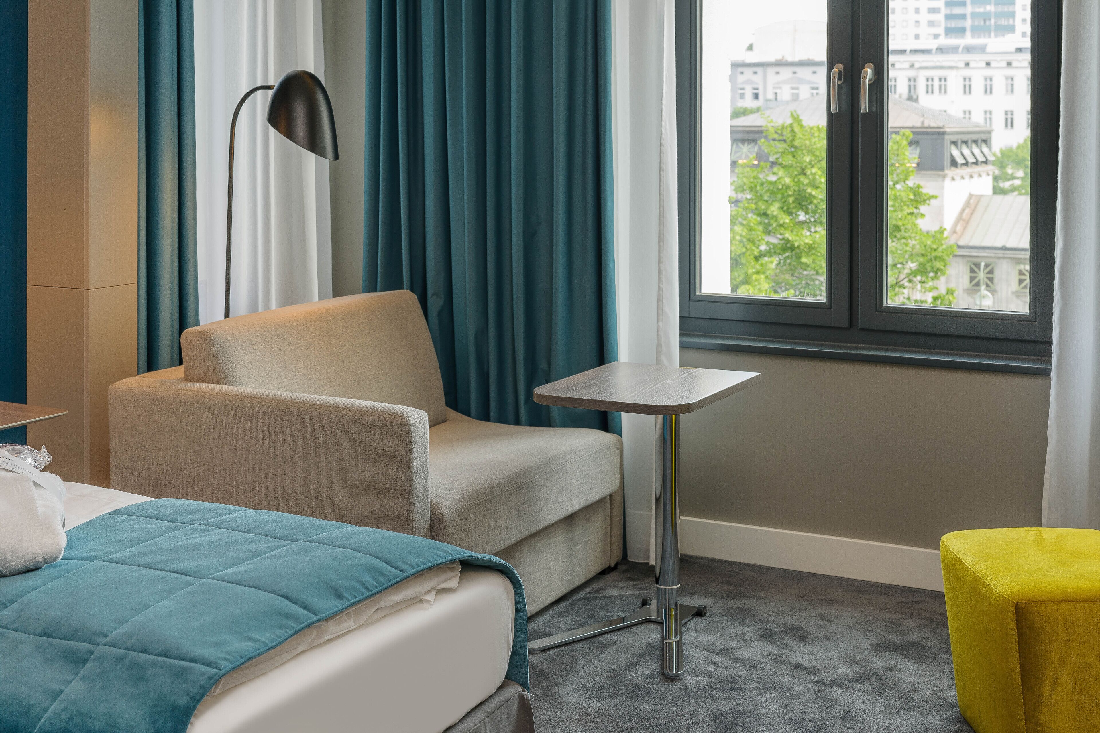 A hotel room with a bed, a chair, a small table, and a window with a city view.