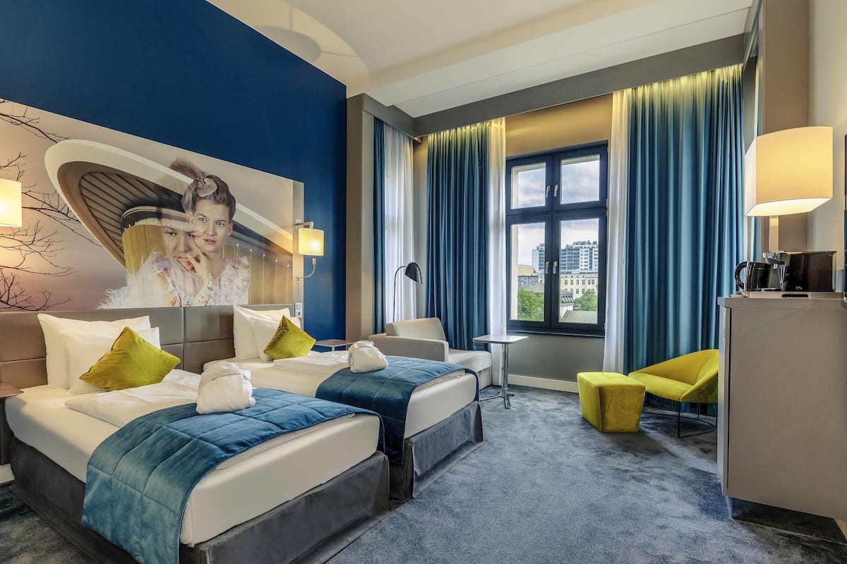 Ein Hotelzimmer mit zwei Betten, einem Schreibtisch, einem Sessel und einem großen Fenster mit Vorhängen.