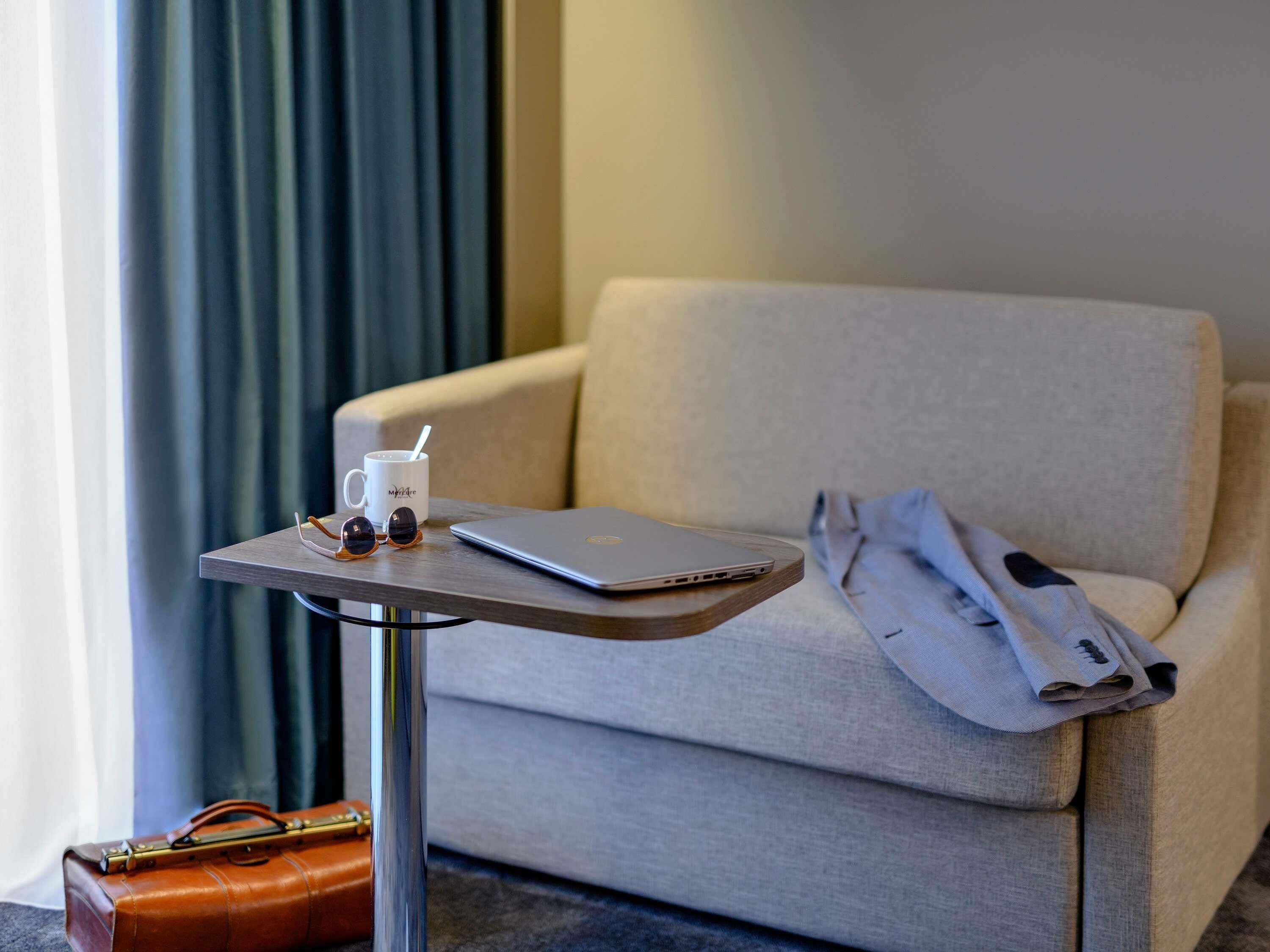 A hotel room with a sofa, a small table with a laptop, a cup, and a pair of sunglasses.