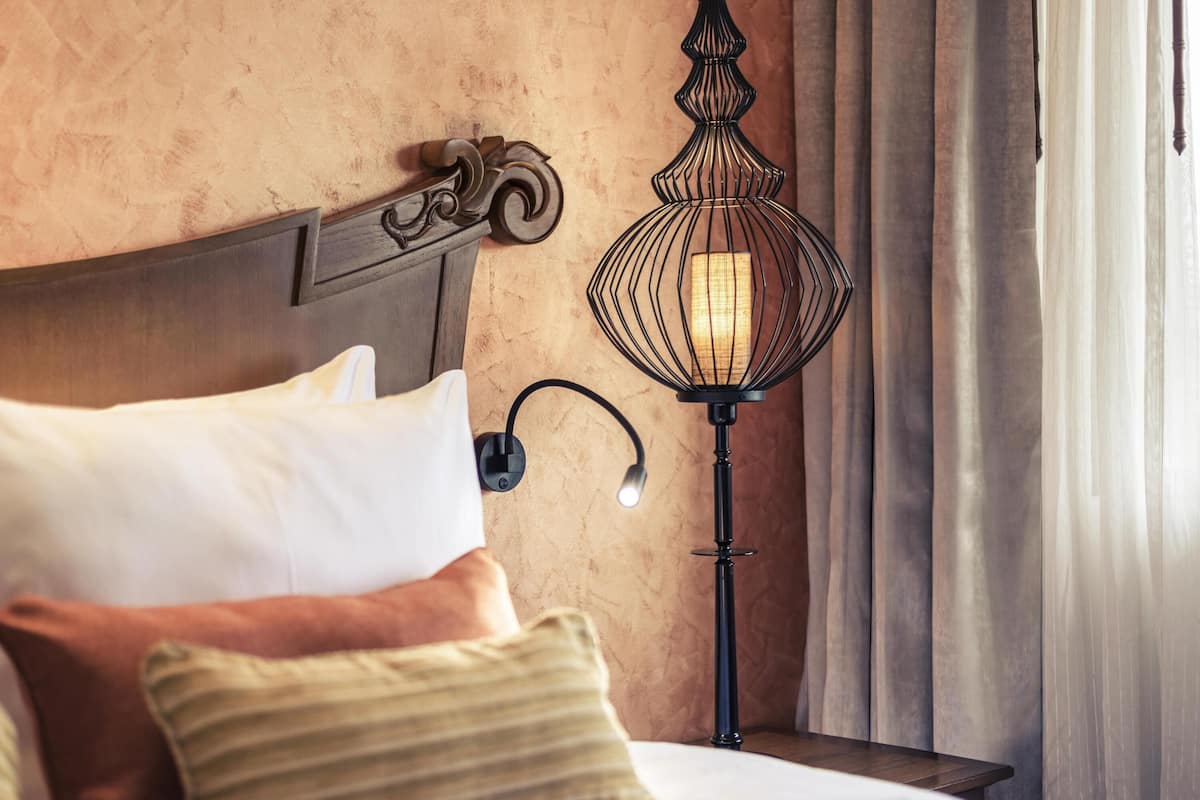 A neatly made bed with pillows, a wall-mounted lamp, and a wooden headboard.