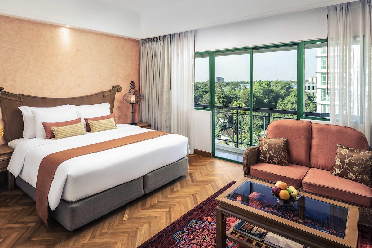 A hotel room with a large bed, a sofa, a coffee table, and a view of greenery and buildings.