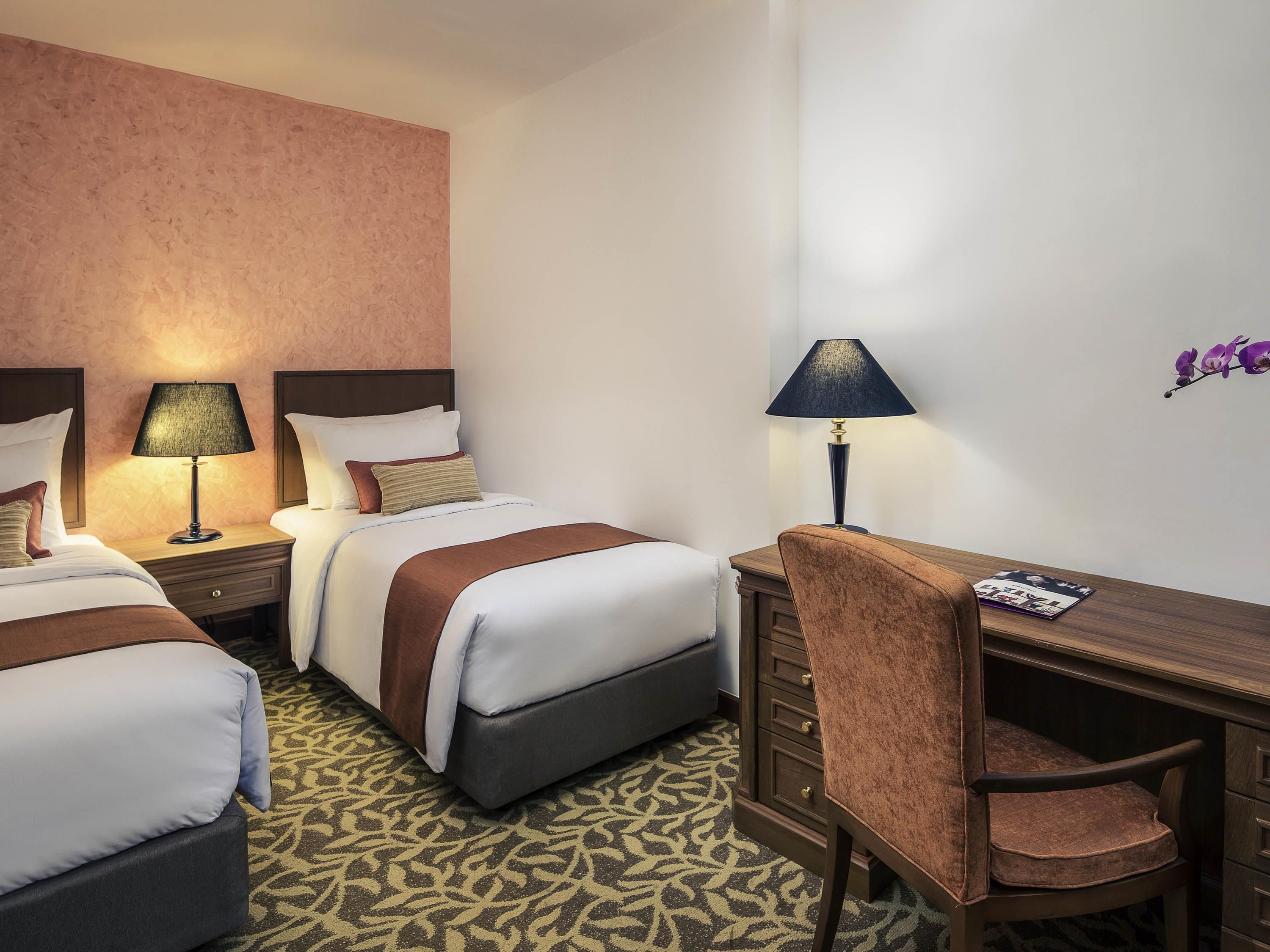 A hotel room with two beds, a desk, a chair, and a wall-mounted lamp.