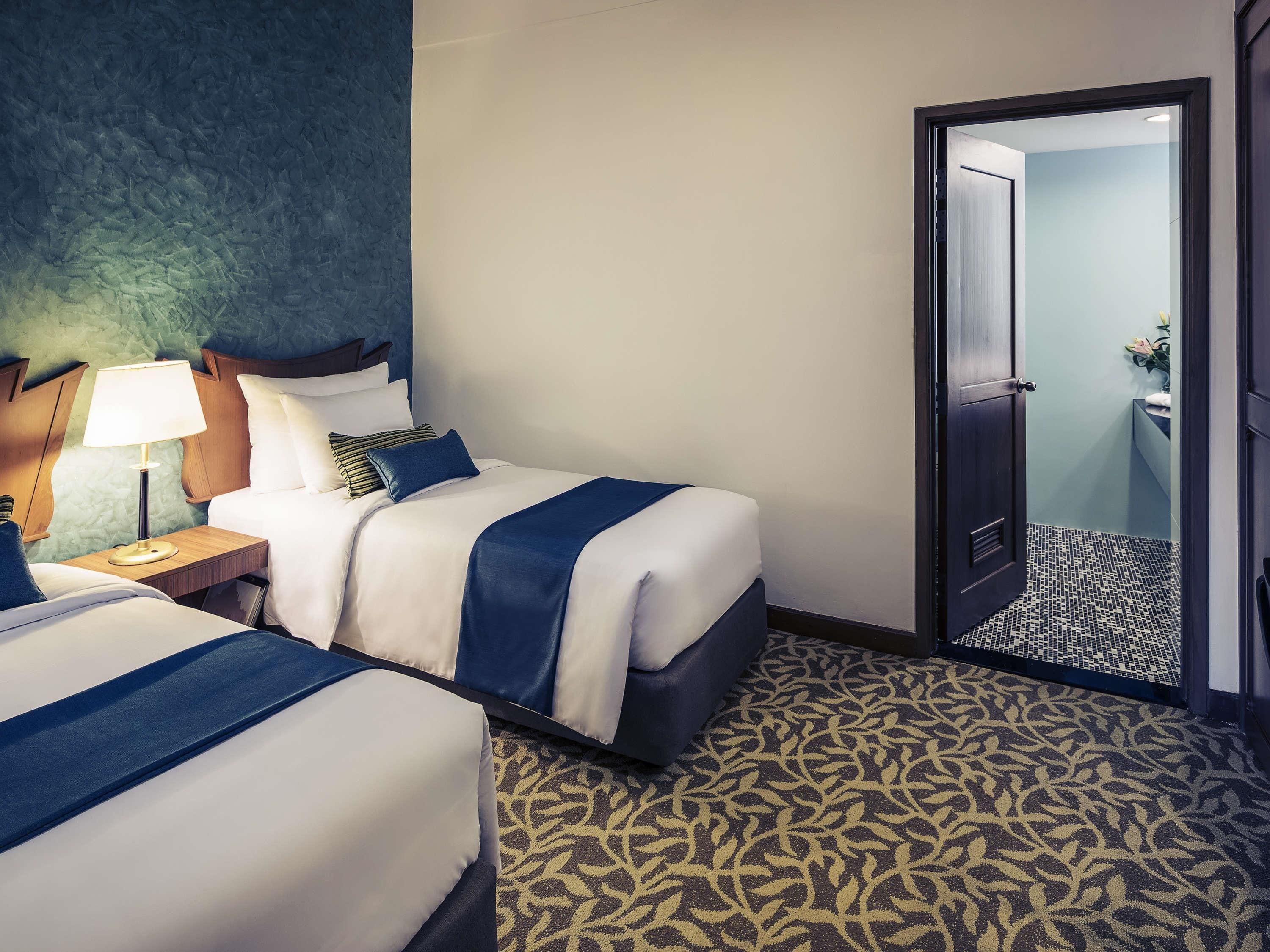 Habitación de hotel con dos camas, una mesita de noche con lámpara, y un baño visible a través de una puerta abierta.
