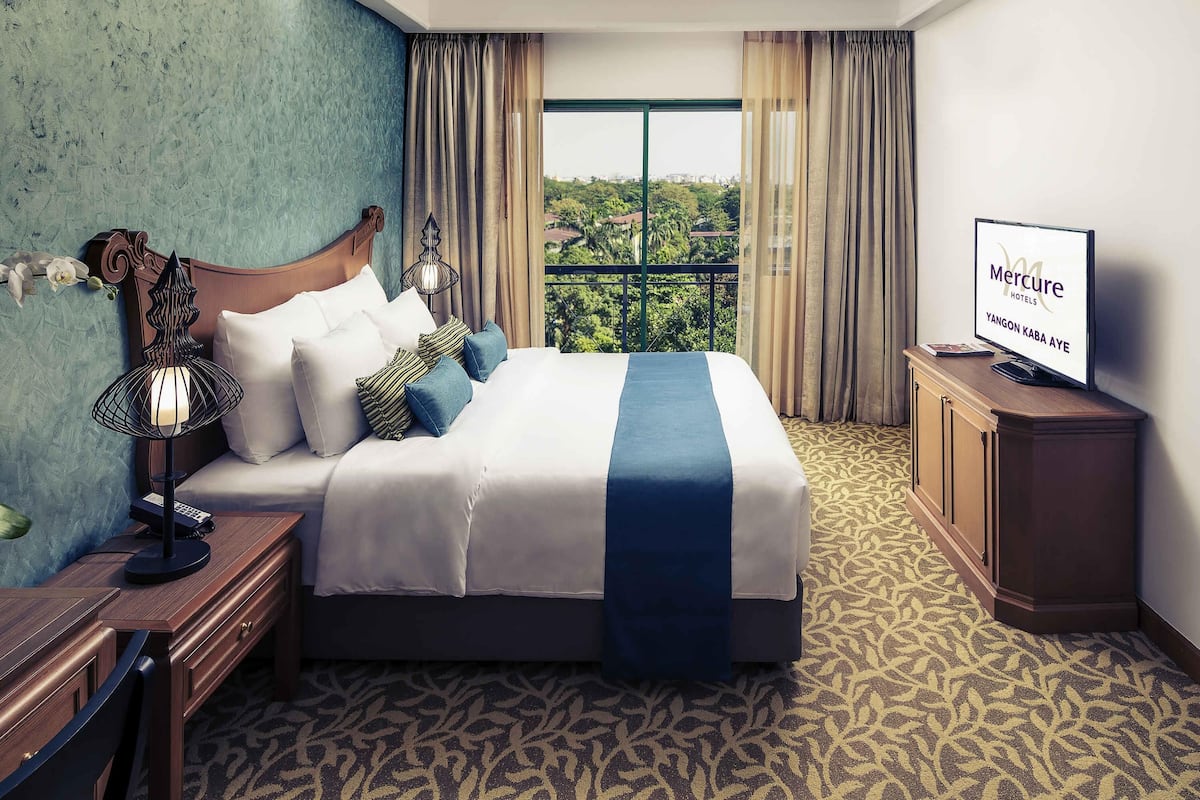 A hotel room with a bed, a TV, a desk with a lamp, and a view of greenery through the window.