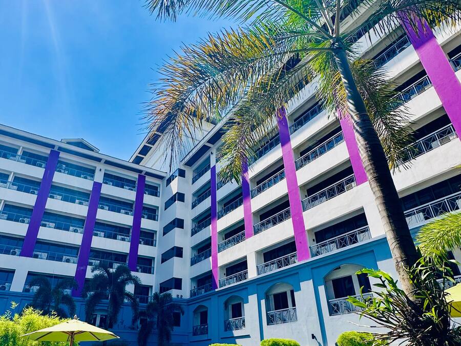 Un hotel con piscina, palmeras y un edificio de varios pisos con detalles morados.