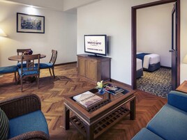 Habitación de hotel con piso de madera tipo espiga, un televisor sobre un mueble de madera, una zona de comedor con mesa y sillas redondas, y una cama con cabecera azul.