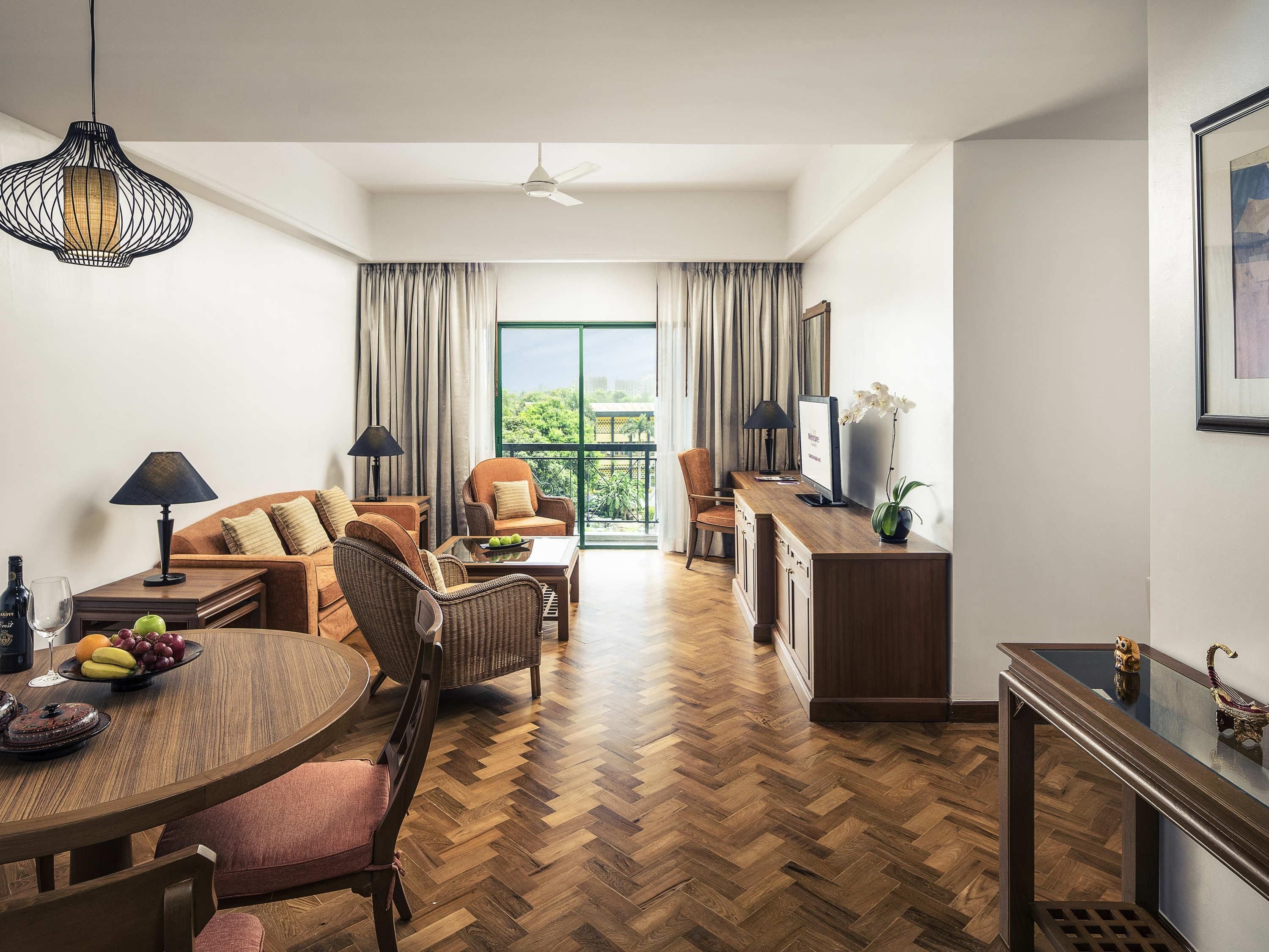 Un salón espacioso con suelo de parqué tipo espiga, una mesa de comedor con fruta, un sofá, un televisor y un balcón con cortinas.