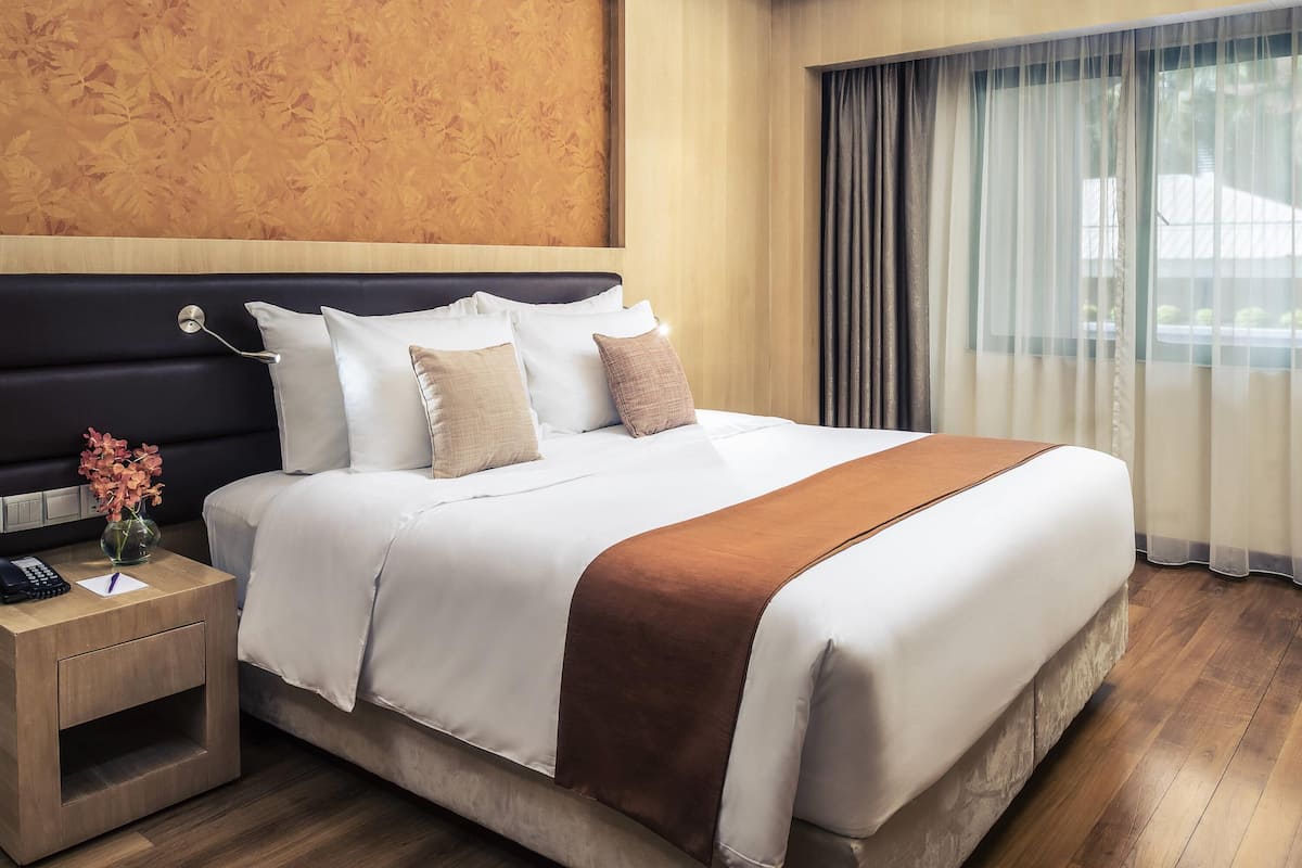A hotel room with a large bed, a bedside table with a phone, a vase with flowers, and a view of the outdoors through sheer curtains.