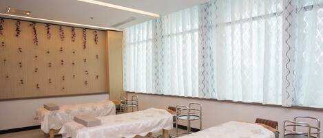 A room with two beds, each with a wooden headboard and a white bedspread. There are two metal chairs and a metal cart with shelves. The room has a wooden panel wall with floral decorations and a large window with patterned curtains.