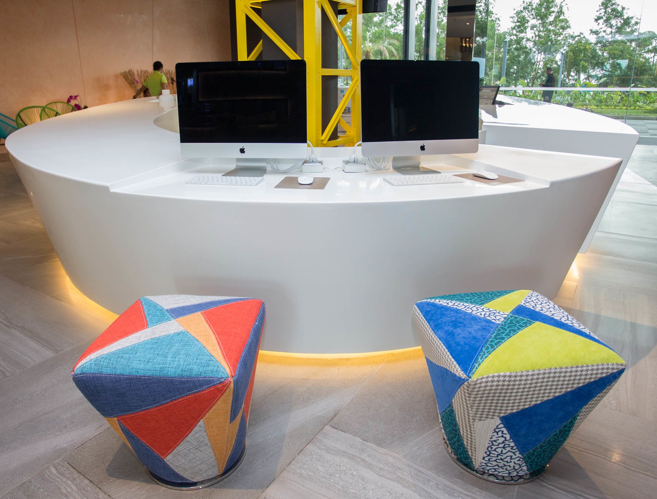 Un comptoir d’accueil équipé de deux ordinateurs Apple iMac, de deux tabourets géométriques aux couleurs vives et, en arrière-plan, d’une structure métallique jaune.