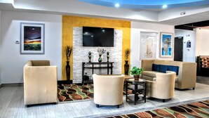 A modern lobby with a flat-screen TV mounted on a stone wall, beige sofas, a patterned rug, and a small table with a plant.