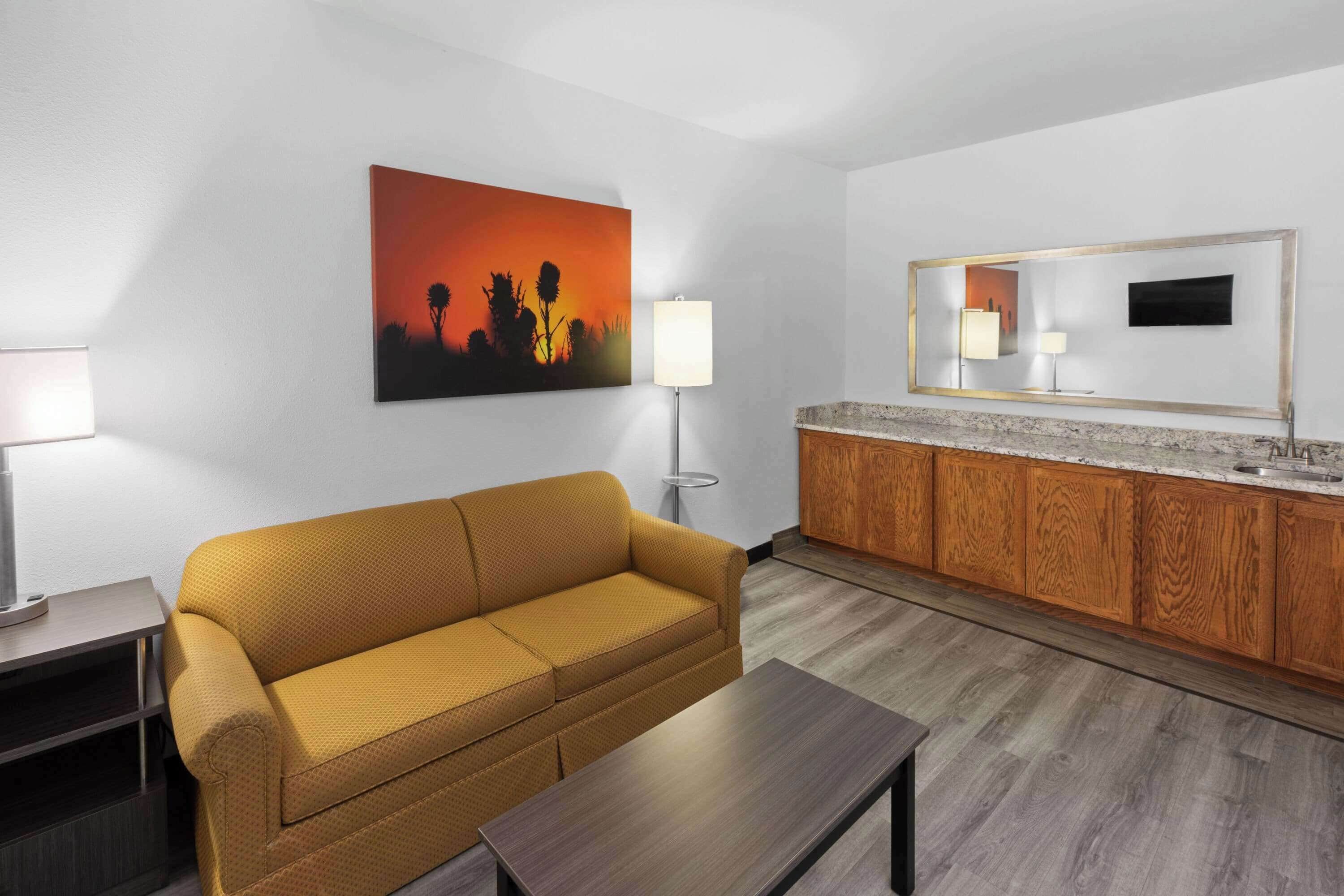 A modern hotel room with a sofa, a coffee table, a painting, a lamp, a mirror, and a wooden cabinet.