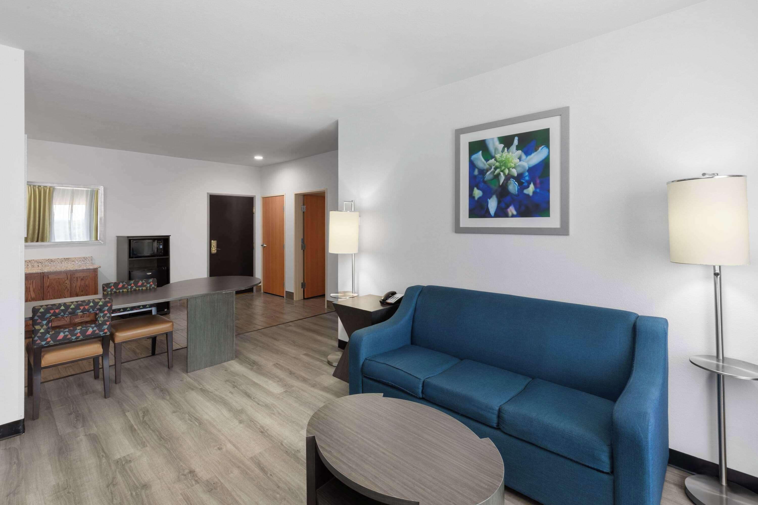 Hotelkamer met een blauwe bank, een ronde salontafel, een zithoek met stoelen, een keuken met een koelkast, en een schilderij aan de muur.