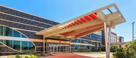 A modern hotel entrance with a covered walkway, large glass windows, and a clear sky.