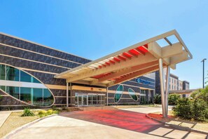 A modern hotel entrance with a covered walkway, large glass windows, and a clear sky.