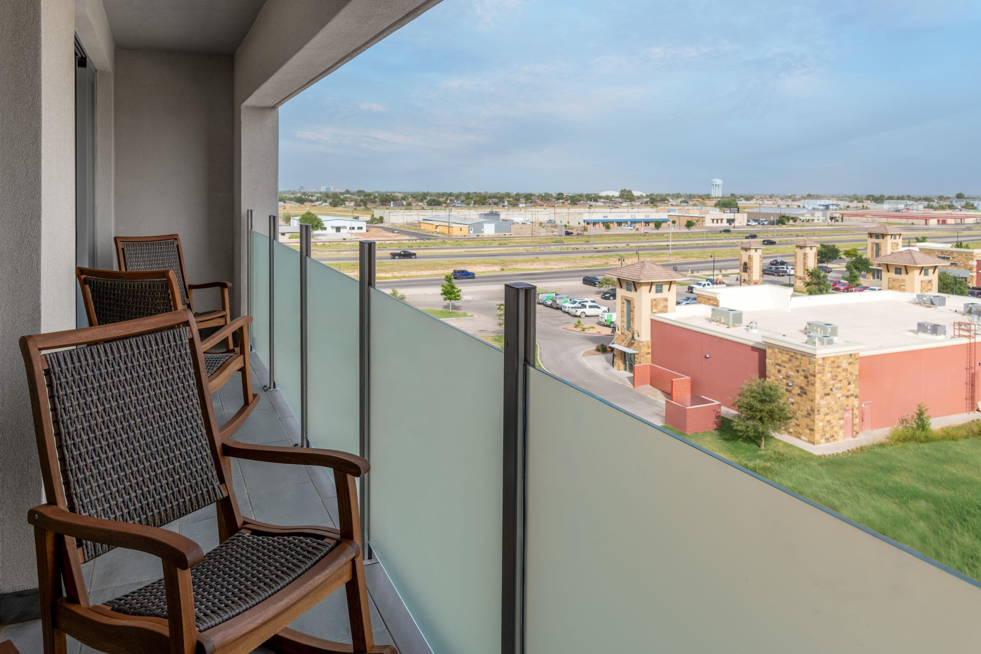 A balcony with two wicker chairs, a view of a parking lot, and buildings in the distance.