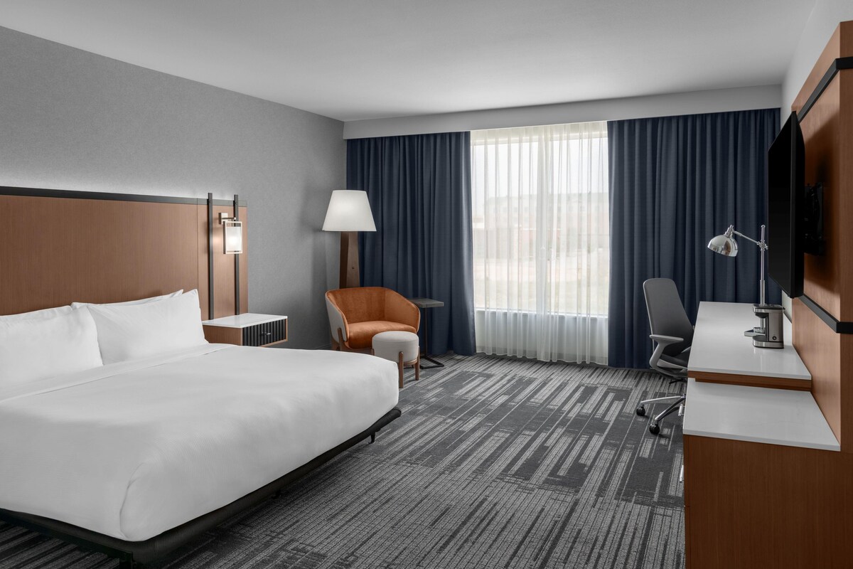 A hotel room with a large bed, a desk, a chair, a lamp, and a TV.