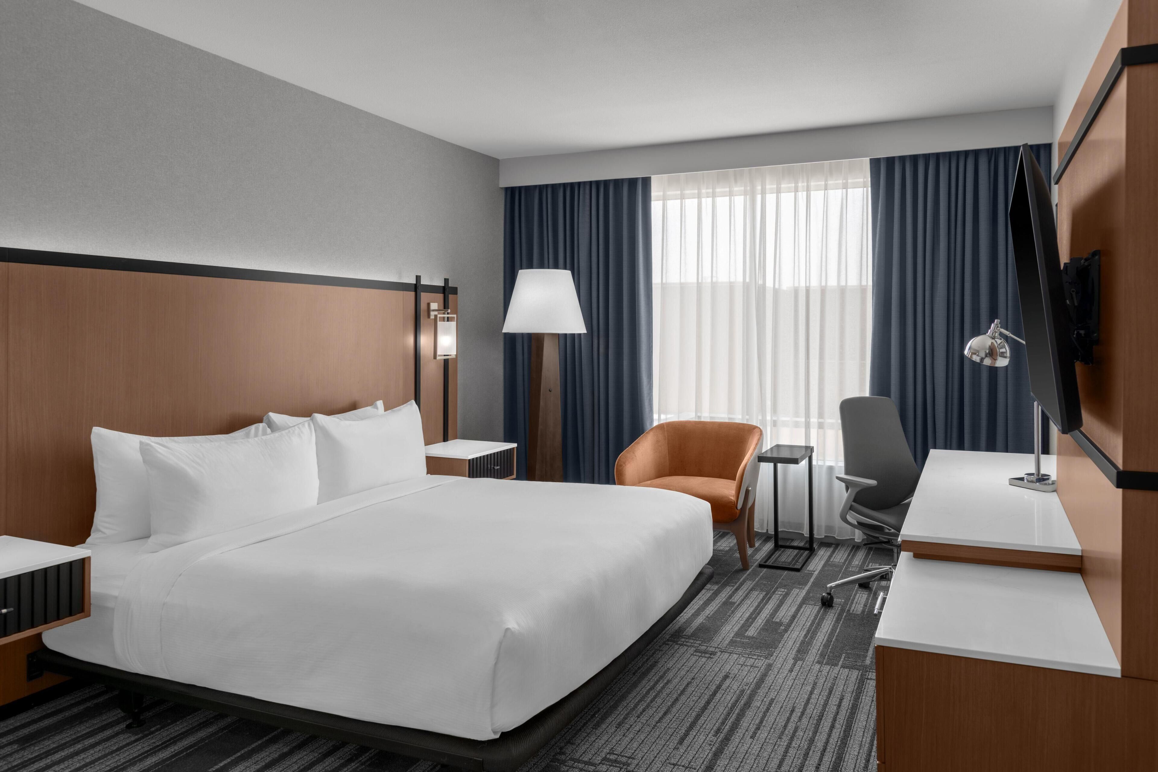 Habitación de hotel con una cama grande, un escritorio, una silla, un televisor y una ventana con cortinas.