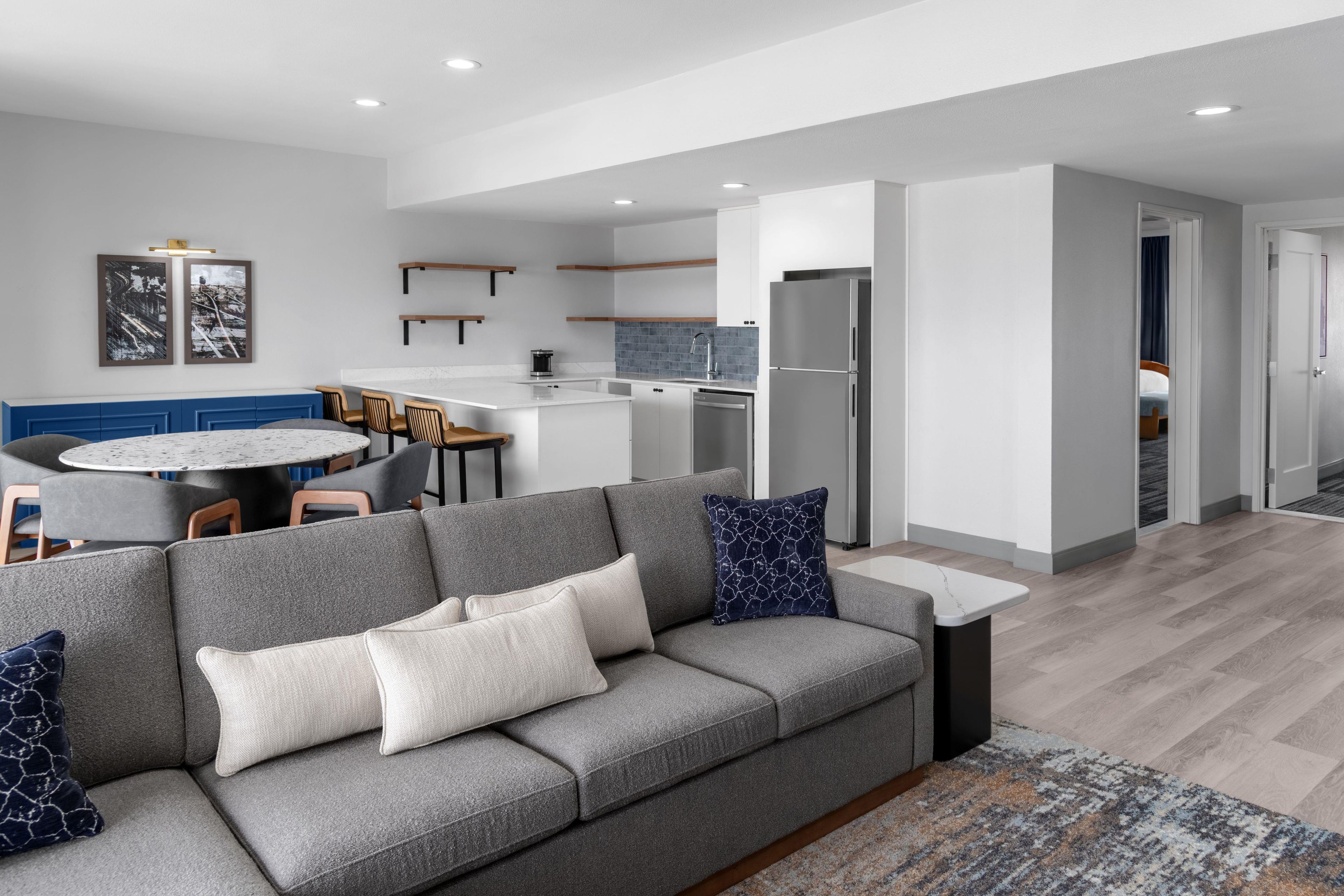 A modern living room with a grey sofa, a round dining table, and a kitchen area with a refrigerator and a coffee machine.