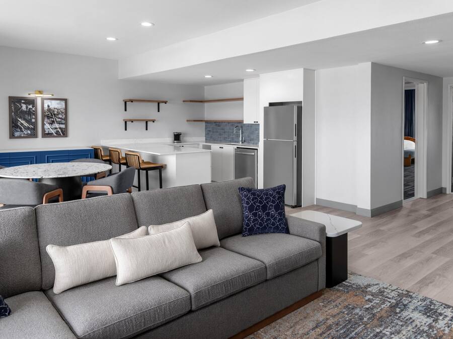 A modern living room with a grey sofa, a round dining table, and a kitchen area with a refrigerator and a coffee machine.