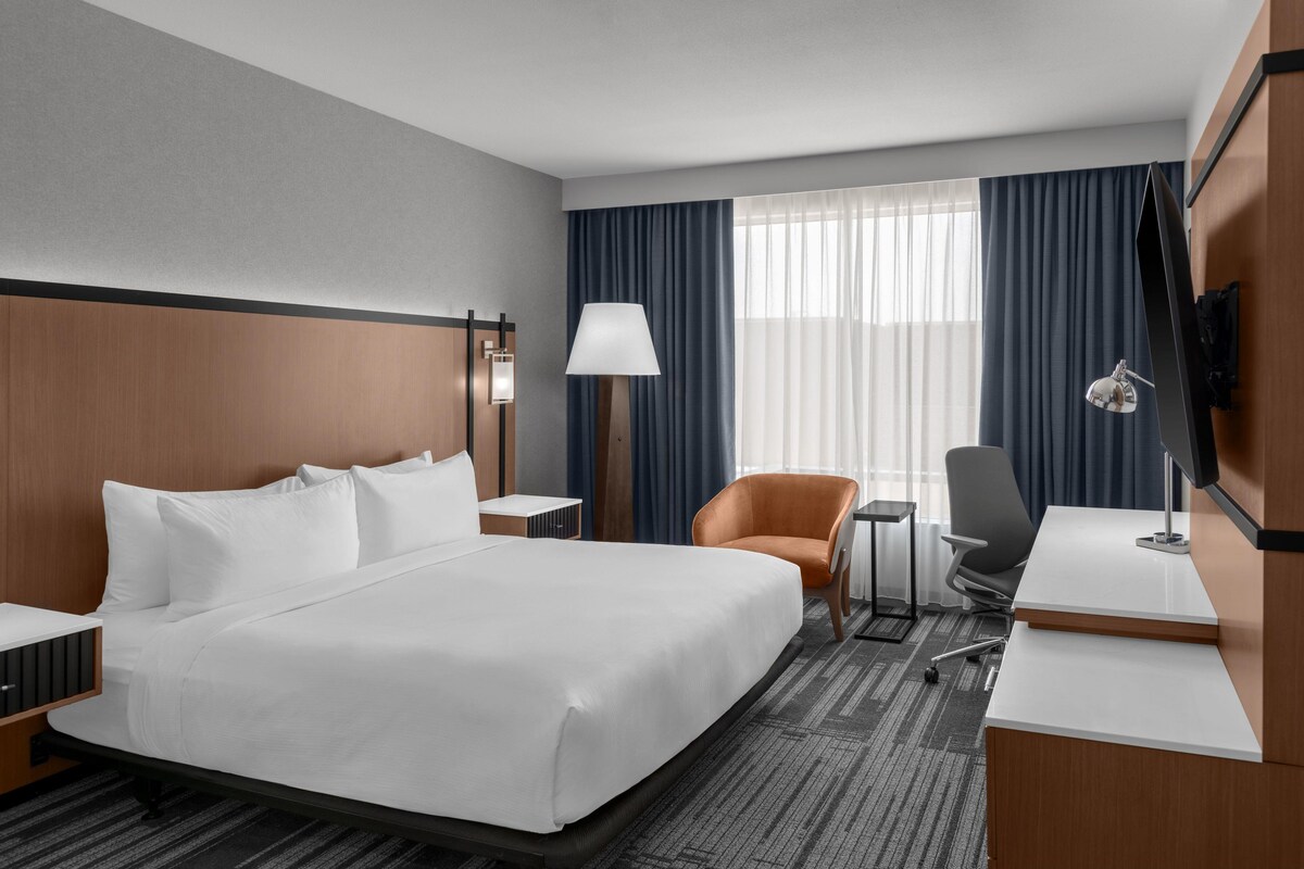 A hotel room with a large bed, a desk, a chair, a TV, and a window with curtains.