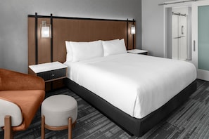 A modern hotel room with a large bed, a nightstand, a seating area with a chair and ottoman, and a wardrobe.