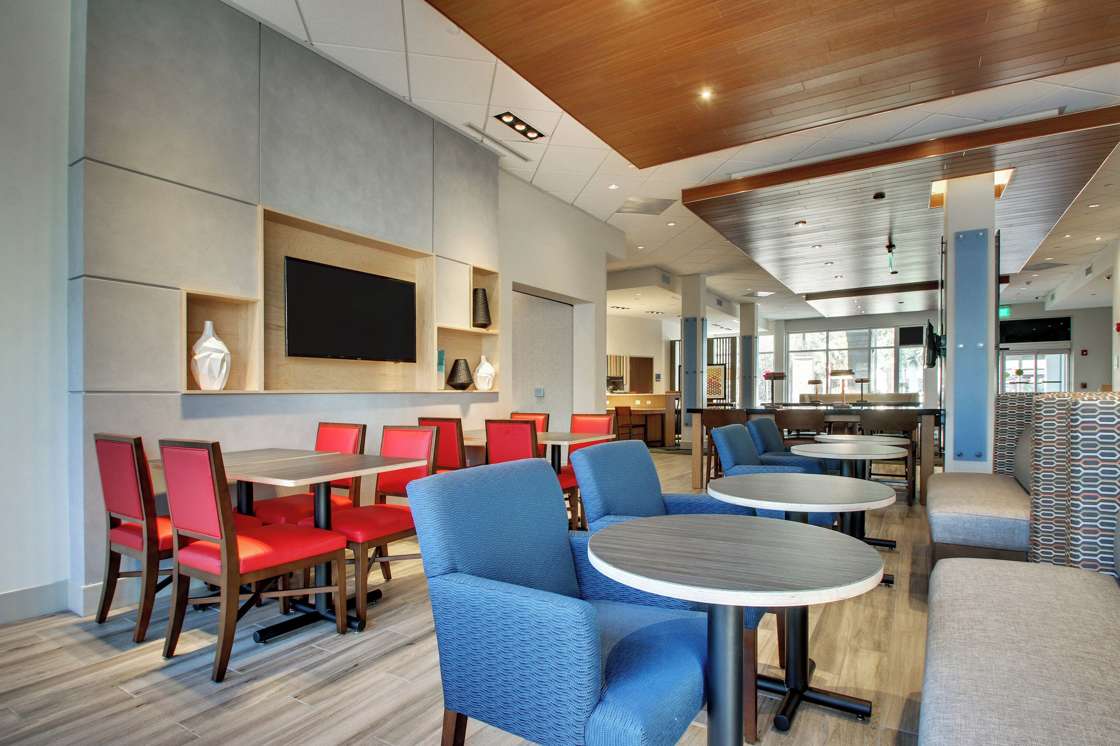 A modern dining area with red and blue seating, a flat-screen TV, and a kitchen in the background.