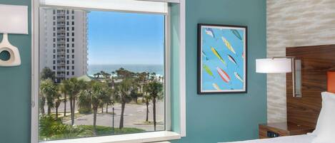 A hotel room with a large window offering a view of palm trees and a building. There is a framed picture on the wall and a bedside table with a lamp.