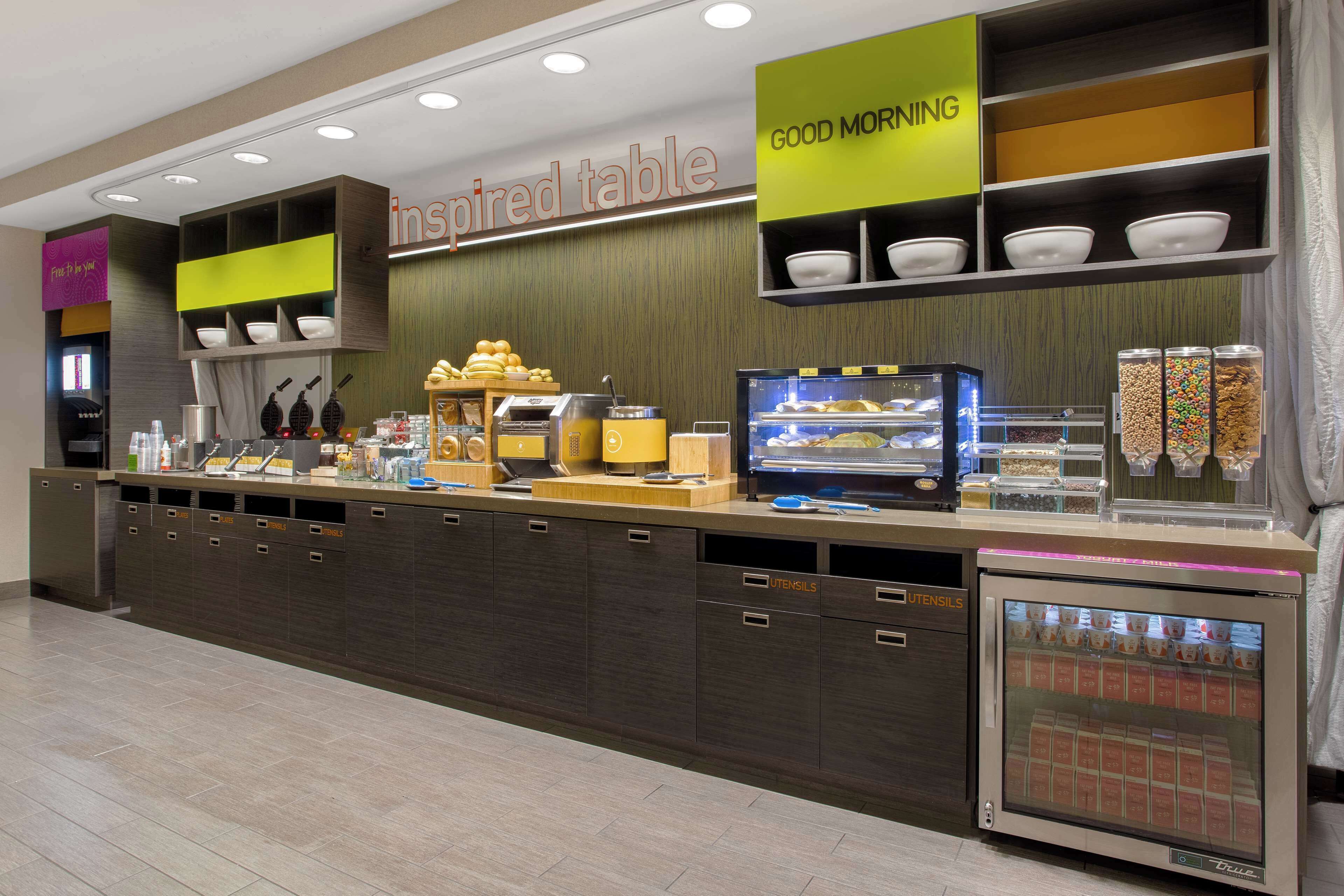 A breakfast bar with a variety of food items and utensils, a refrigerated display case, and a sign for 'Good Morning'.