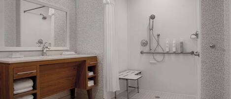A modern bathroom with a wooden vanity, a large mirror, a shower with a handheld showerhead, and a small stool.