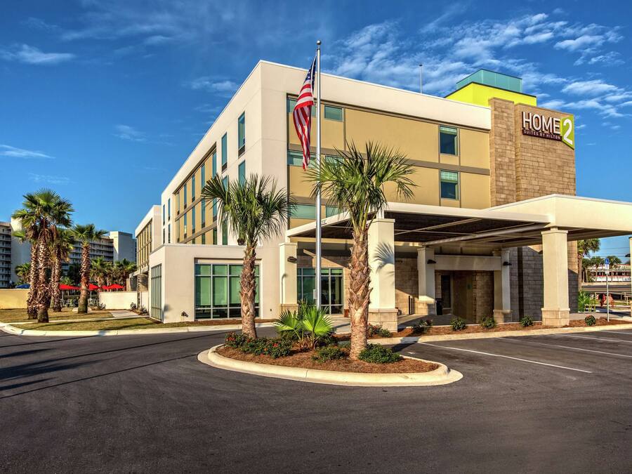 Un hotel moderno con una destacada bandera estadounidense, un letrero que dice "Home 2 Suites by Hilton" y un área de acceso cubierta con palmeras.