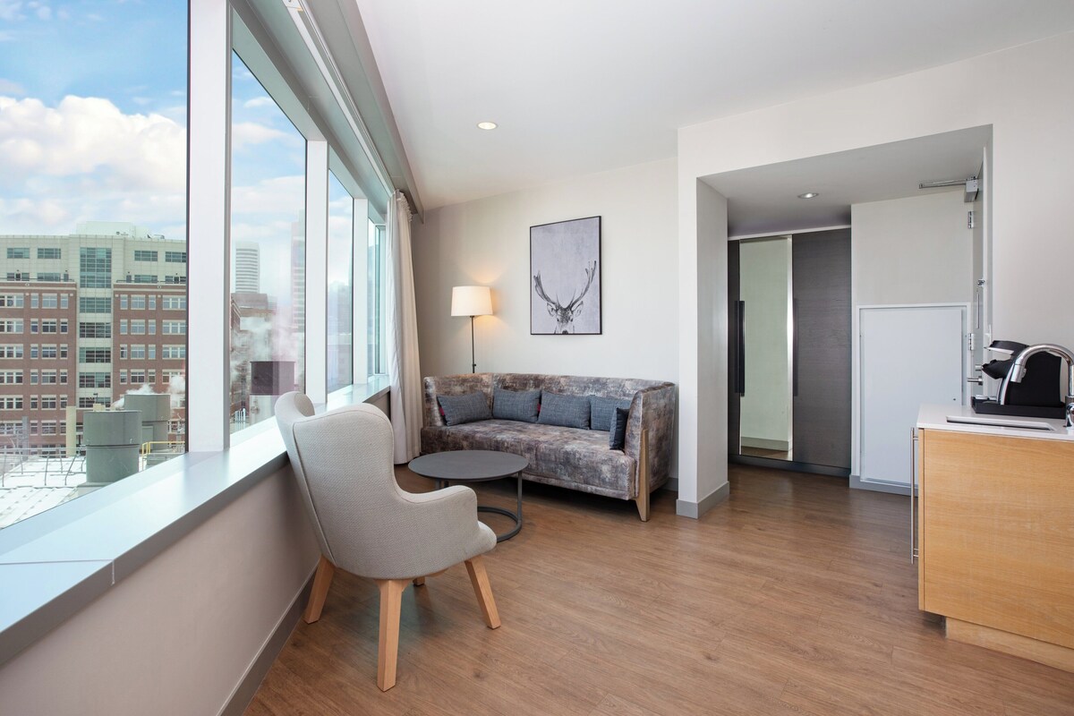 A modern living room with a sofa, armchair, and coffee table. A large window offers a view of buildings outside.
