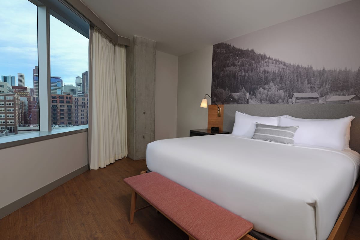 A hotel room with a large bed, a bench, a nightstand, a lamp, and a cityscape view through the window.