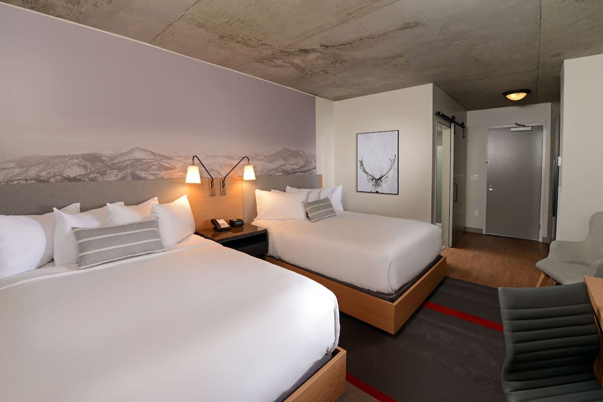 A hotel room with two beds, a desk, a chair, a wall-mounted lamp, and a door.
