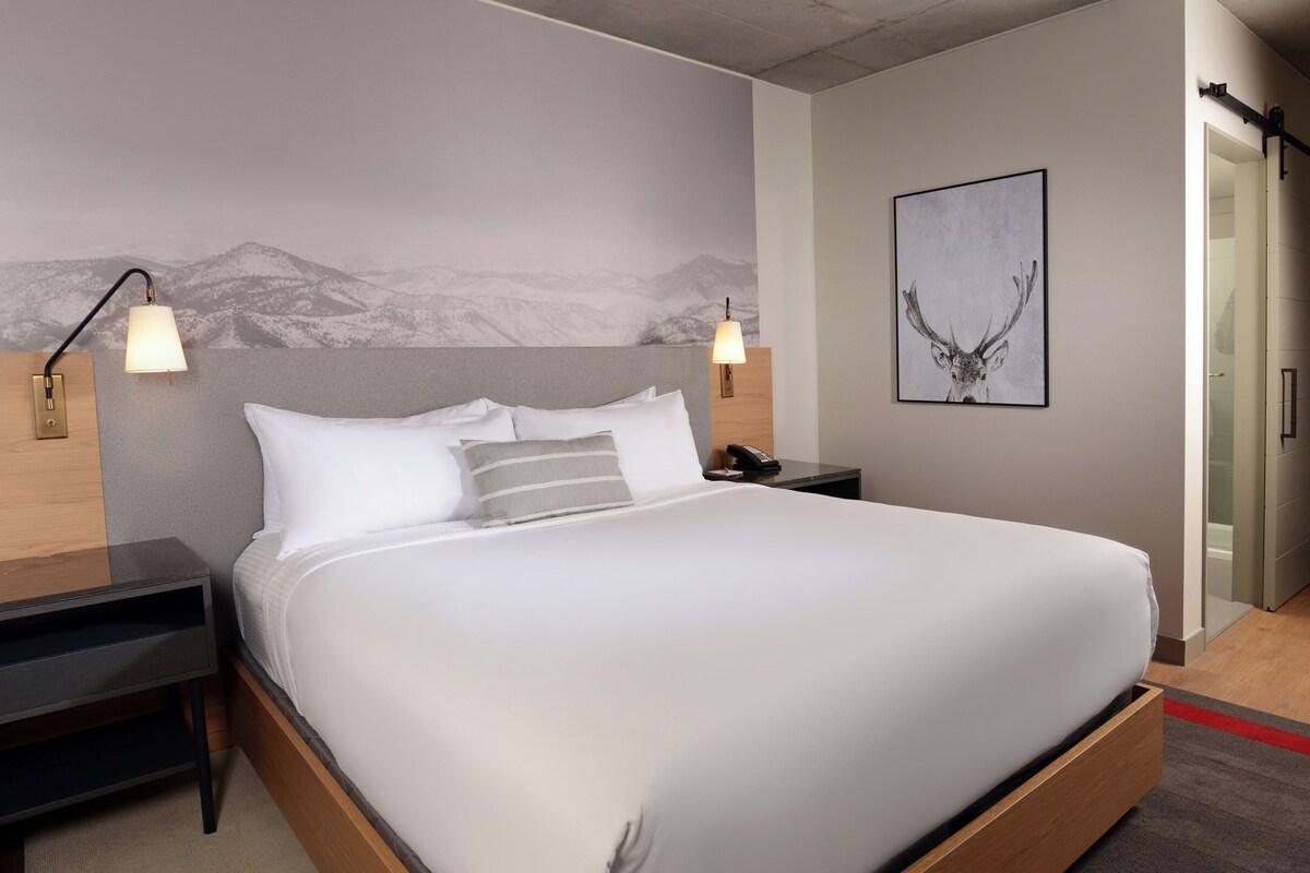 A modern hotel room with a large bed, two bedside tables, a nightstand, a wall-mounted lamp, a framed picture, and a door leading to a bathroom.