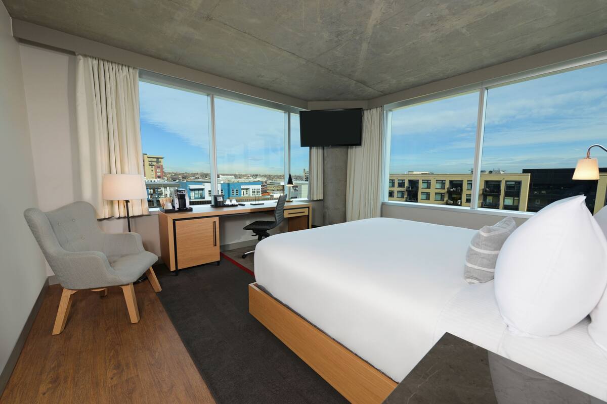A hotel room with a large bed, a desk with a chair, a TV mounted on the wall, and a view of the city through large windows.