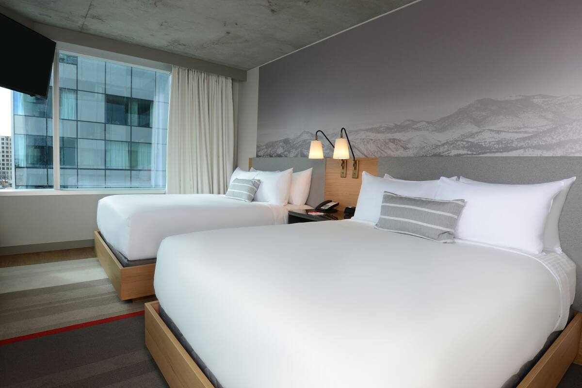 A hotel room with two beds, a large window with curtains, a wall-mounted phone, and a mountain view mural.
