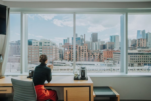 A person seated at a desk with a coffee machine, facing a large window with a cityscape view.