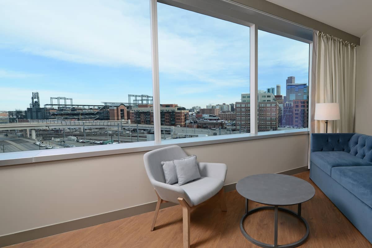 A modern hotel room with a large window offering a city view, a grey armchair, a round table, and a blue sofa.