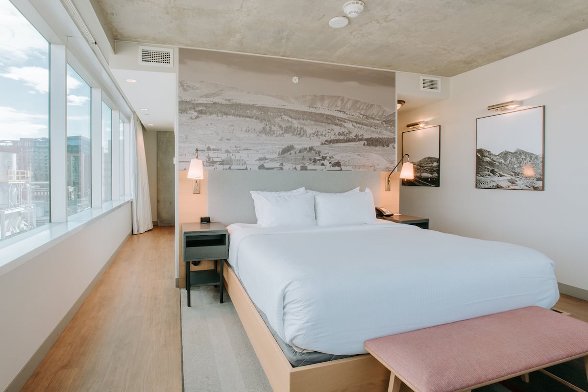 A modern hotel room with a large bed, a bench, a nightstand, and a wall-mounted lamp.