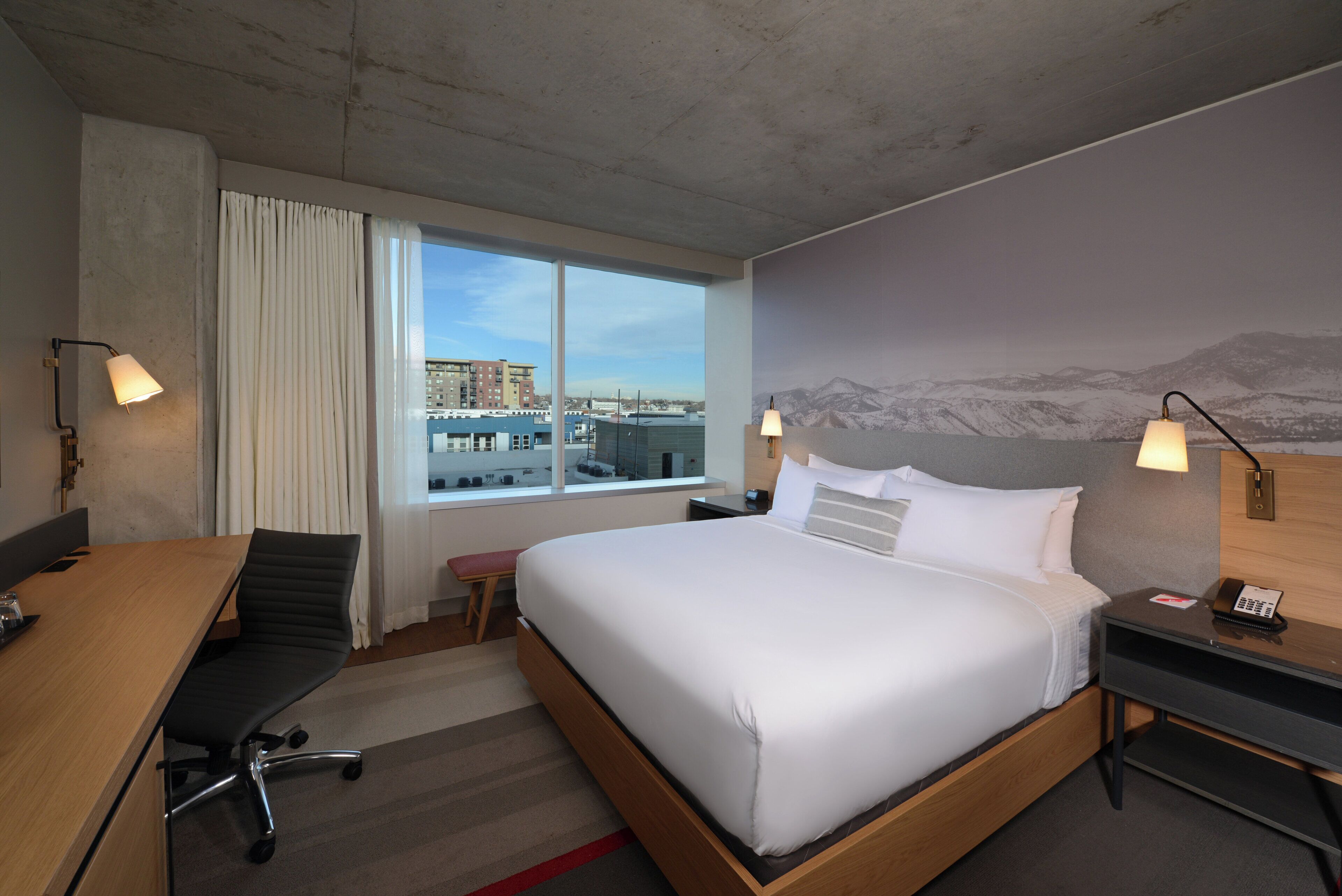 A hotel room with a large bed, a desk, a chair, a nightstand with a phone, and a window with a city view.