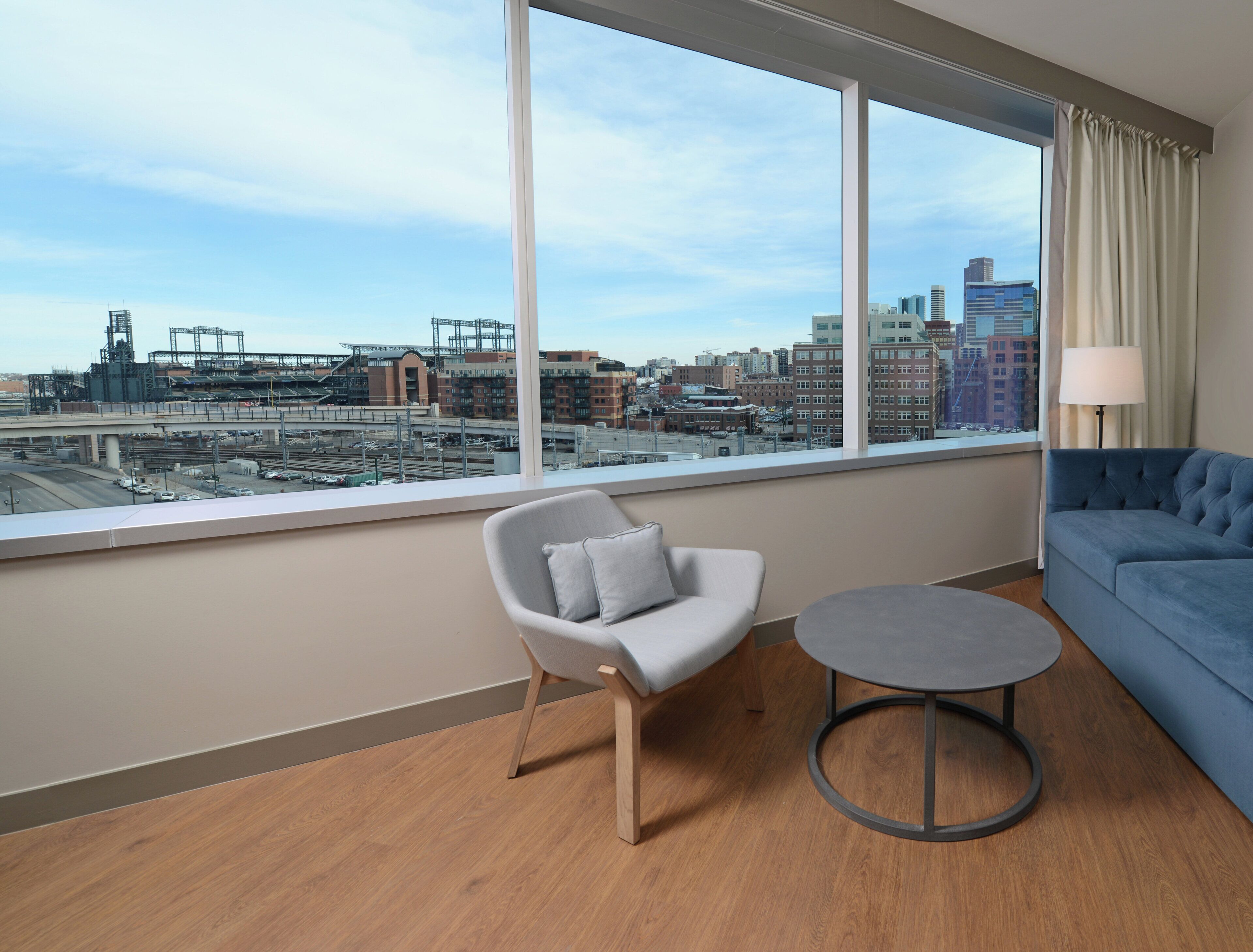 A modern hotel room with a large window offering a city view, a grey armchair, a small round table, and a blue sofa.