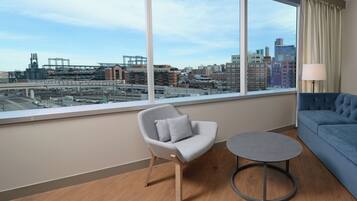 A modern hotel room with a large window offering a city view, a grey armchair, a small round table, and a blue sofa.