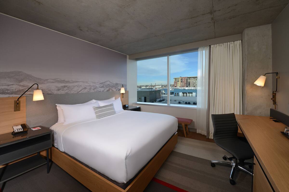 A hotel room with a large bed, a desk, a chair, a phone, and a window with a city view.