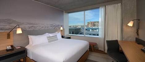 A hotel room with a large bed, a desk, a chair, a phone, and a window with a city view.