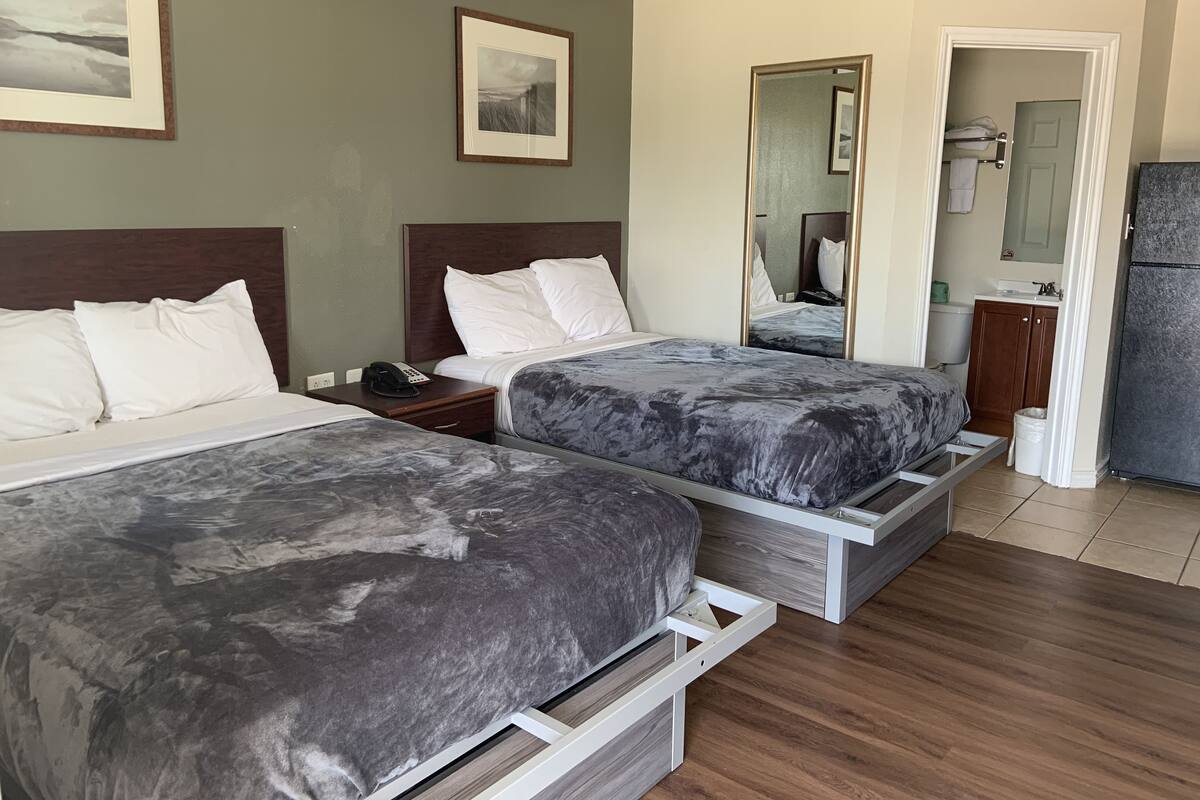 A hotel room with two beds, a wooden floor, a bathroom with a mirror, and a refrigerator.