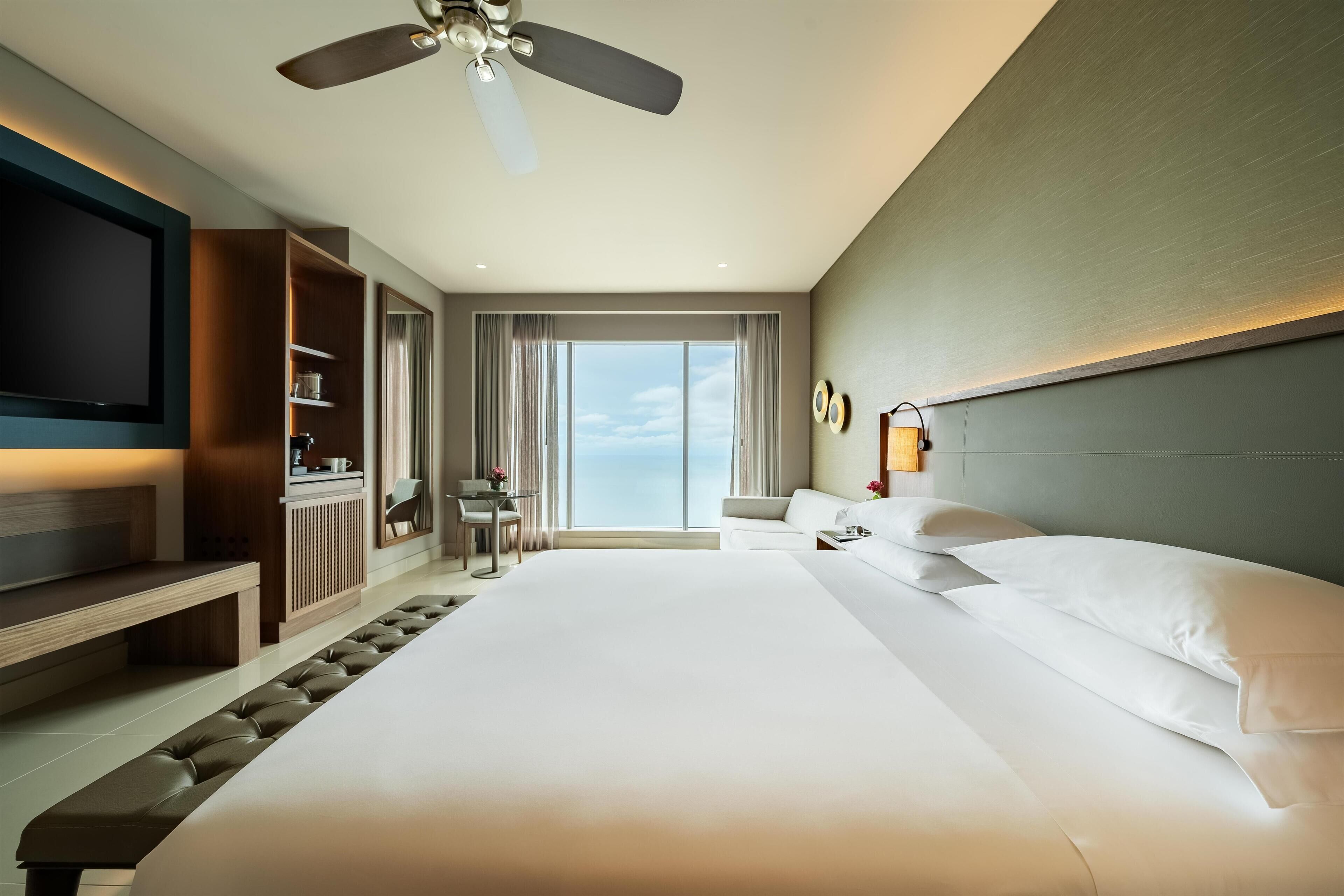 Una habitación de hotel moderna con una cama grande, un televisor de pantalla plana, un ventilador de techo, un área de descanso con una mesa y vistas al cielo a través de amplios ventanales.